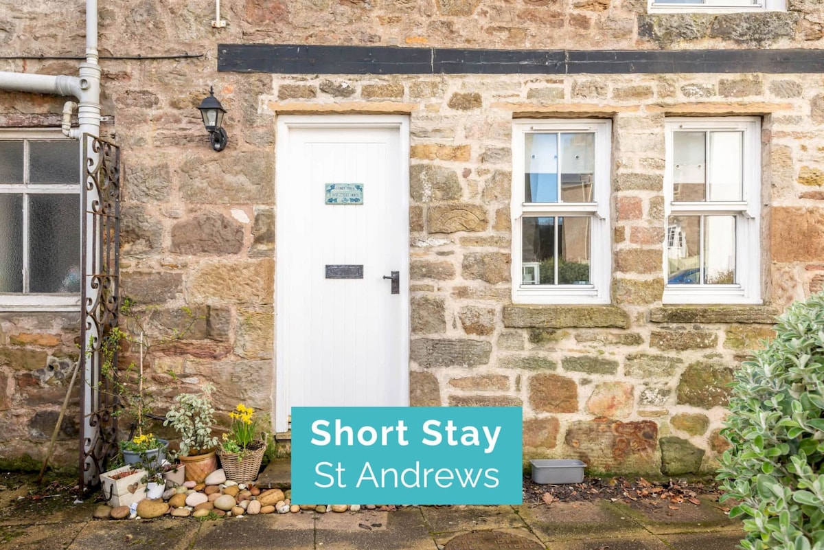 The entrance to the cottage features a compact white door framed by natural stone walls. Two windows on either side allow natural light, while a small garden bed filled with plants adds a touch of greenery to the inviting facade.