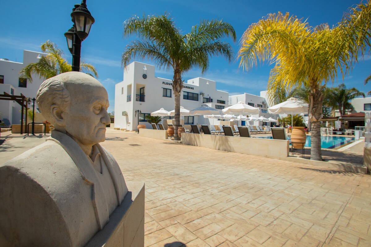 A stone statue is positioned in the foreground, with palm trees and sun loungers visible nearby. A sparkling pool is seen in the background, surrounded by a sun terrace and white buildings, creating a sunny, inviting atmosphere.