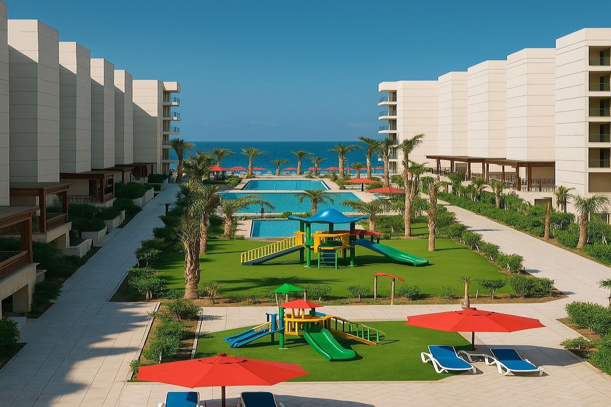 A spacious outdoor area is shown with a green playground featuring colorful slides and play structures. Sun loungers with umbrellas are positioned around a large pool, with palm trees lining the edges. The sea is visible in the background under a clear blue sky.