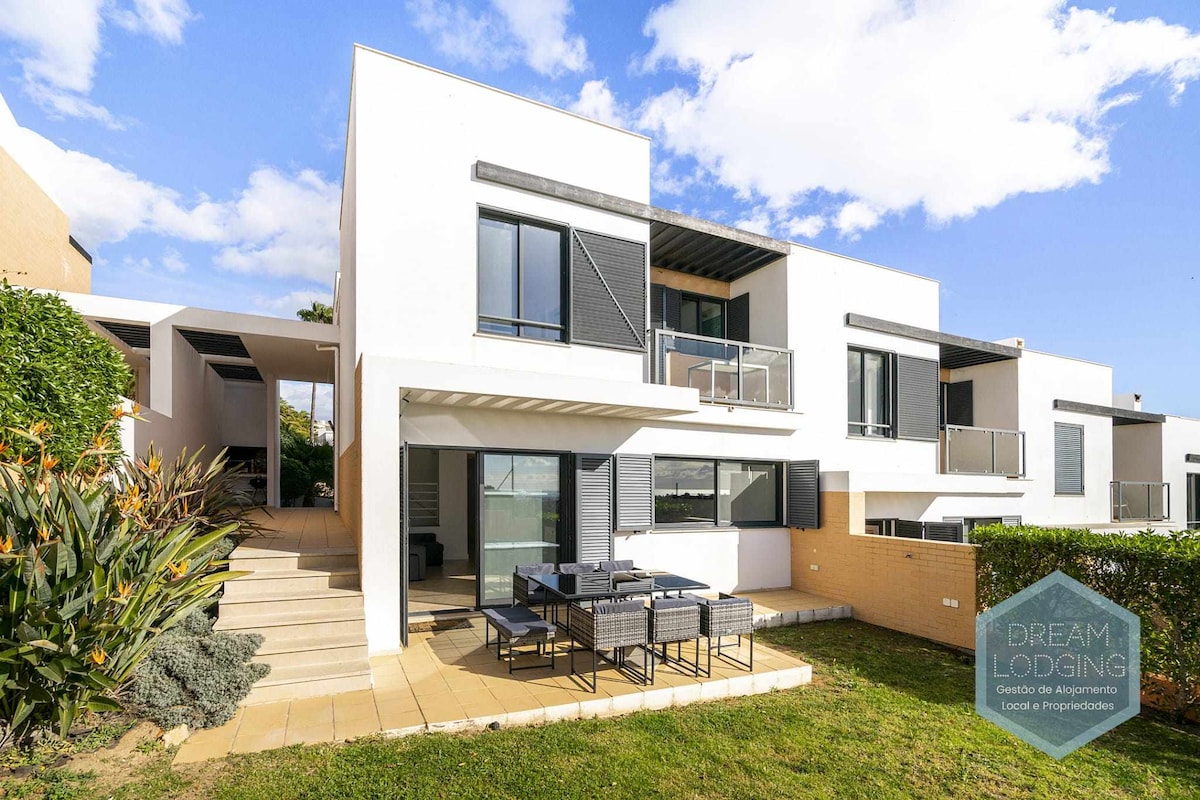 The exterior of a modern two-story property is presented, featuring large balconies and sliding doors. A spacious patio area is equipped with a dining table and chairs, surrounded by lush greenery and well-maintained landscaping. The sky is clear with a few clouds.