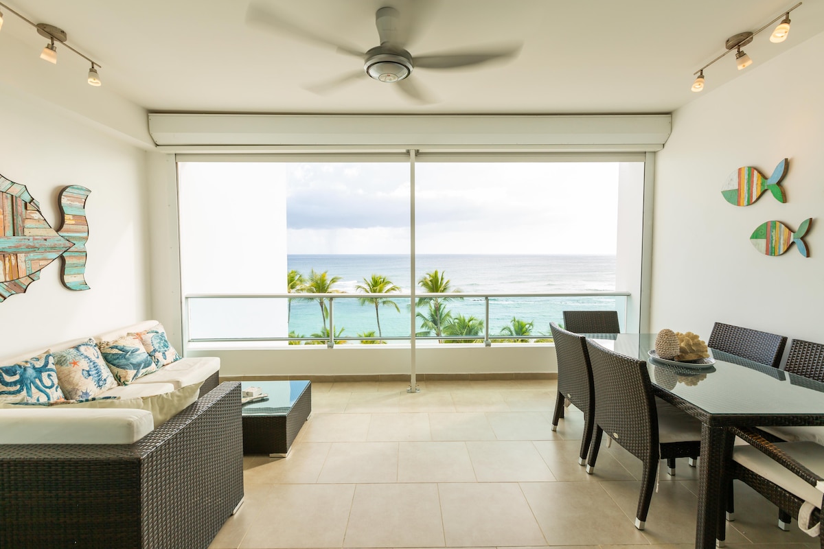 A bright balcony space furnished with a comfortable seating area and a dining table. The ocean view is captured through large glass doors, enhancing the connection with the outdoor scenery. Coastal-themed decor adds a touch of warmth to the inviting atmosphere.
