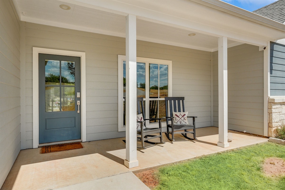 A shaded porch area features two black rocking chairs positioned near a light blue front door. A welcome mat is placed at the entrance, with a grassy area visible nearby and a clear sky overhead.