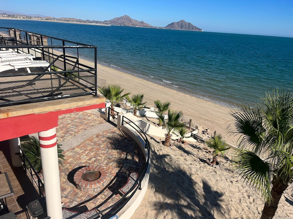 The beachfront patio features a circular seating area surrounded by palm trees. Relaxing lounge chairs are positioned nearby on the sandy shore, providing unobstructed views of the Sea of Cortez and distant mountains. The coastline showcases a tranquil sandy beach extending along the water.