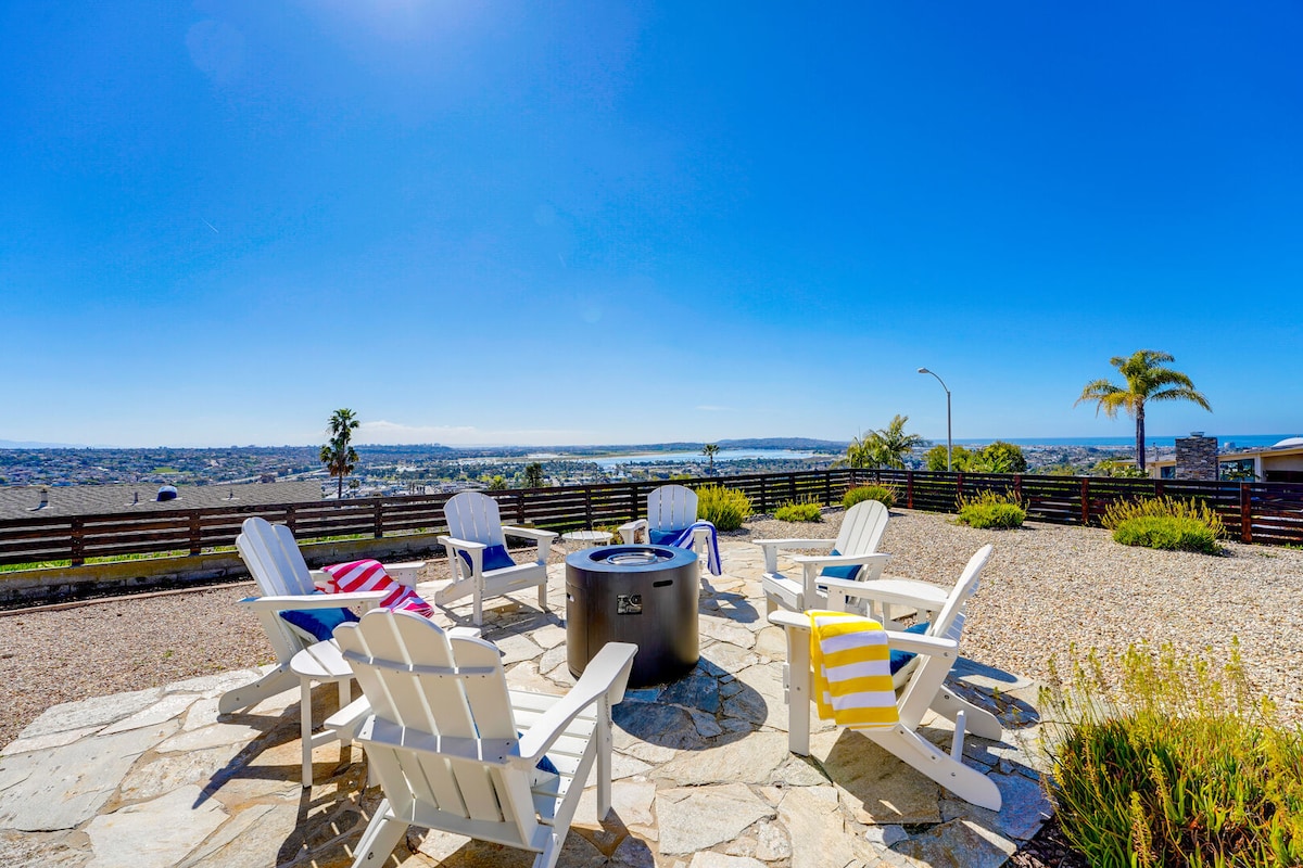 An outdoor seating area features six white Adirondack chairs arranged around a circular fire pit. The patio is paved with stone and framed by landscaping, providing views of the ocean and surrounding area against a bright blue sky.