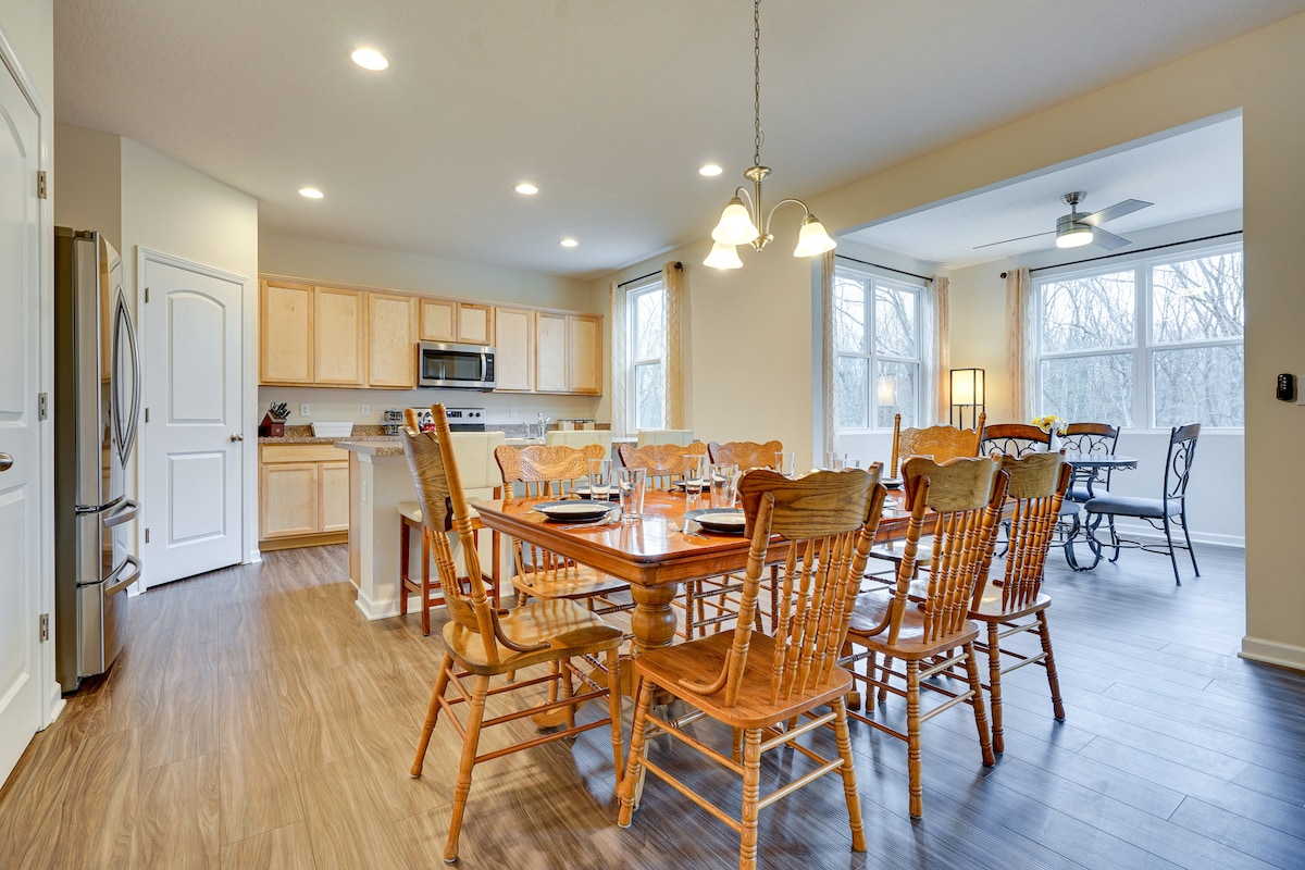 An open dining area is showcased with a large wooden table surrounded by several wooden chairs. A well-equipped kitchen is visible in the background, complemented by large windows that allow natural light to brighten the space.