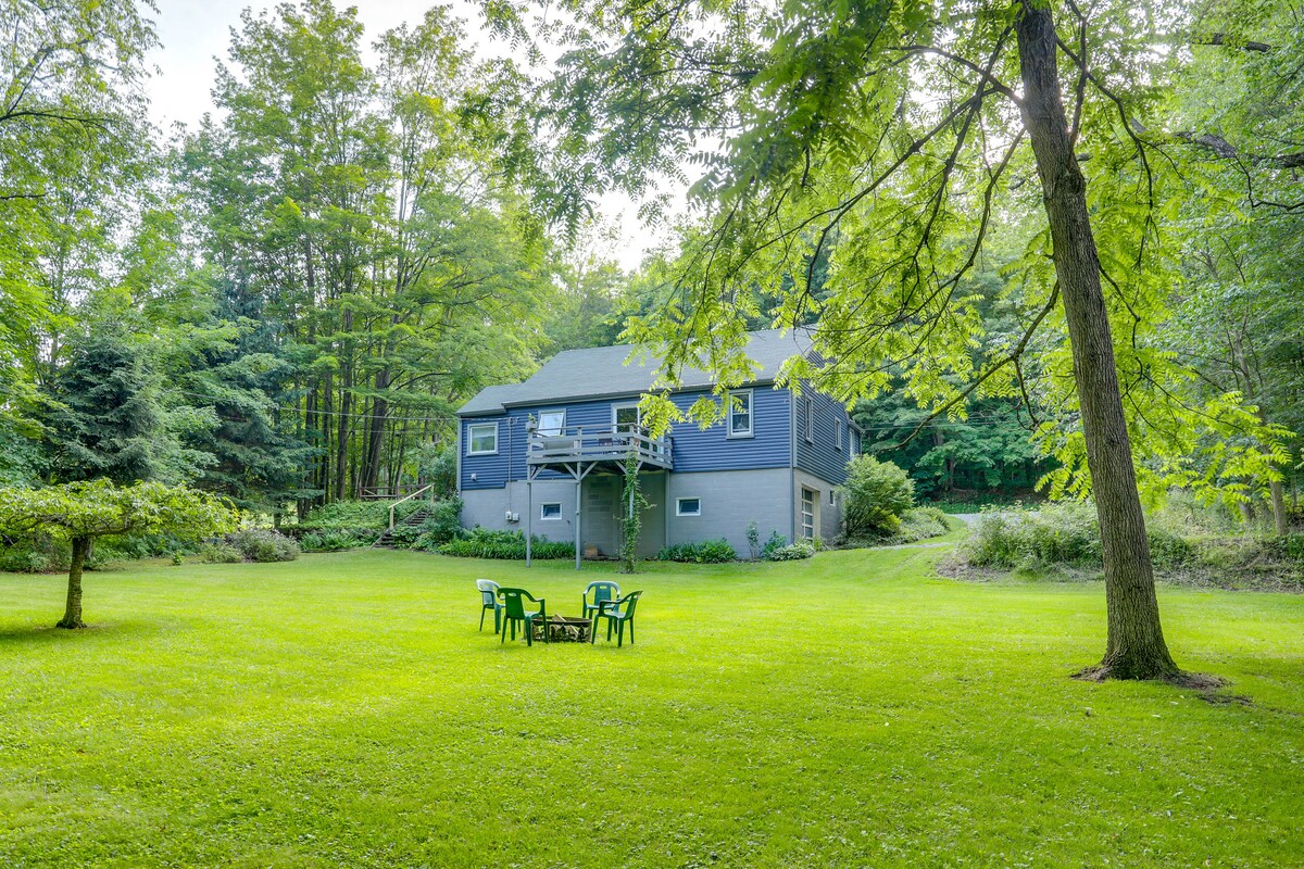 The exterior of the vacation rental is surrounded by lush greenery, featuring a spacious lawn. A seating area consisting of four chairs is positioned near a tree, inviting relaxation among the natural landscape. The house stands with a deck overlooking the serene property.
