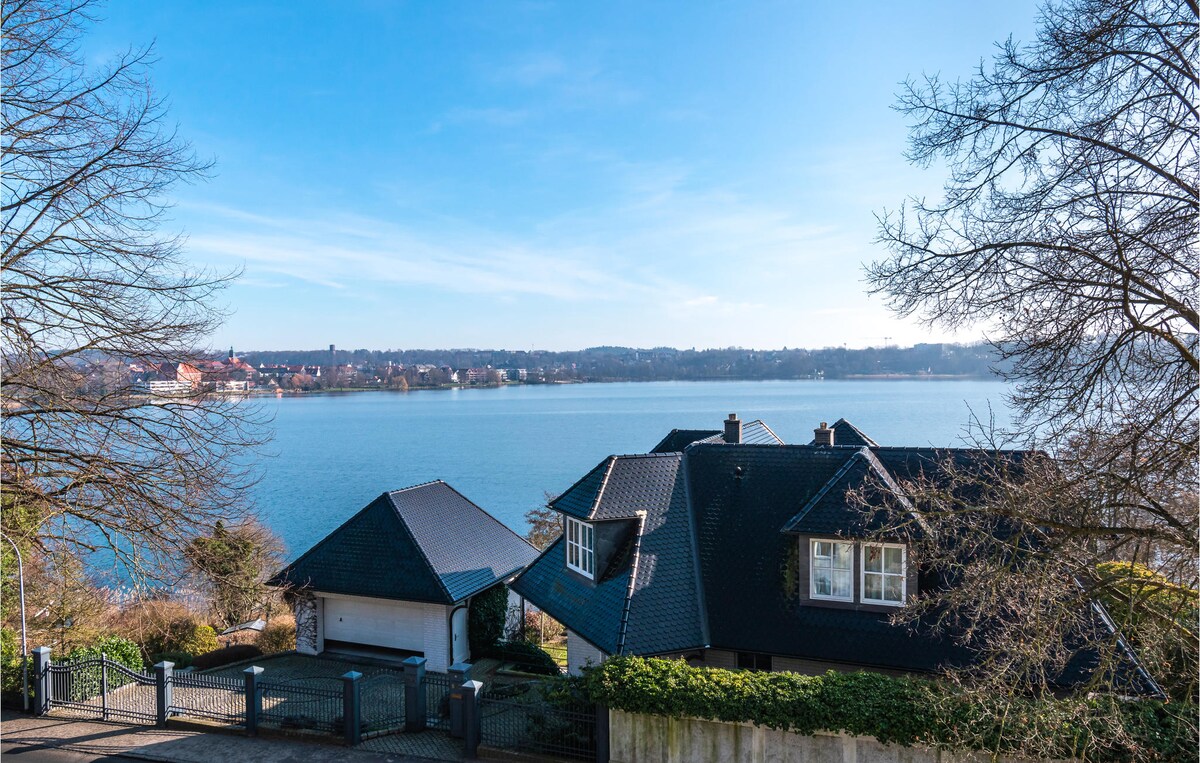 A tranquil view of Lake Ratzeburg is captured, featuring calm waters and a clear blue sky. The image showcases a charming residence with a dark-roofed structure and surrounding greenery, enhancing the scenic lakeside atmosphere.