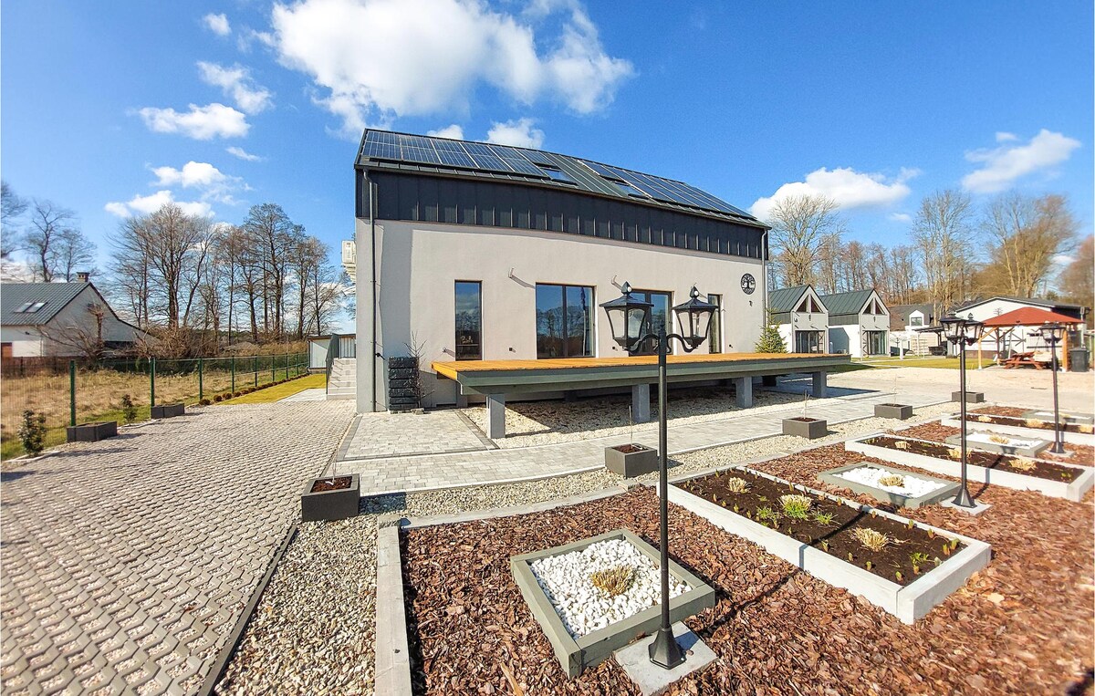 The modern cottage features a flat-roof design with large windows framed by a wooden terrace. The outdoor space includes landscaped areas with gravel and plants. The surrounding environment reveals a mix of greenery and neighboring buildings, under a blue sky dotted with clouds.