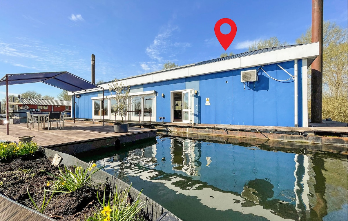 The houseboat's exterior is showcased against a clear blue sky, with bright blue walls reflecting in the calm water. A large terrace with shaded seating is visible, alongside landscaping that includes greenery and a flowerbed near the water's edge.