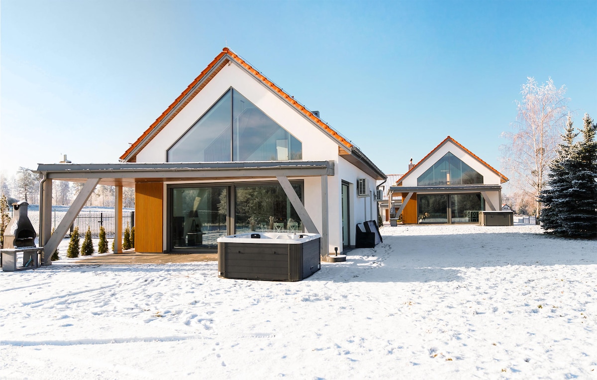 Two modern vacation homes are set against a snowy backdrop, featuring large glass panels that allow ample natural light. A private hot tub is positioned on the deck, complementing the serene outdoor space dotted with trees and snow-covered ground.