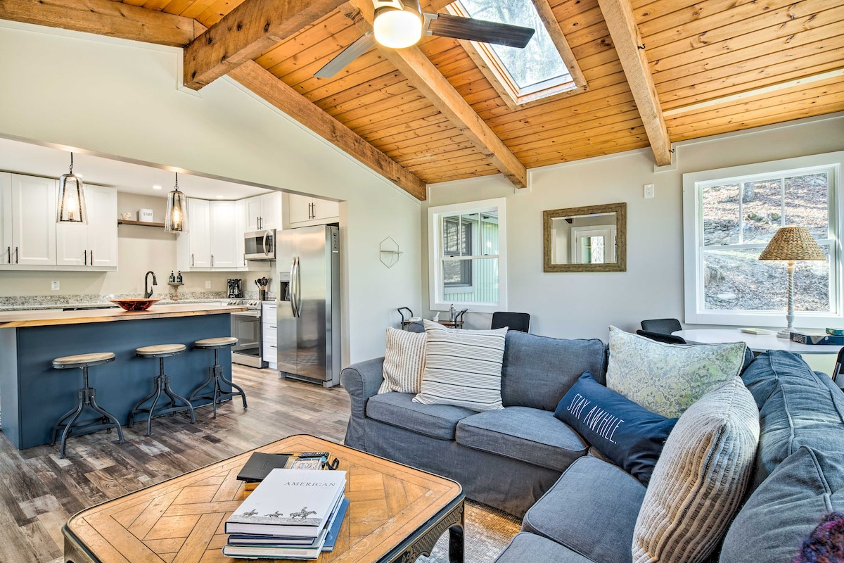 A bright living area features a sectional sofa with a variety of textured throw pillows. Skylights above allow natural light to brighten the space, while the open layout connects to a modern kitchen with bar seating. A rustic coffee table sits atop the wood-floored area.