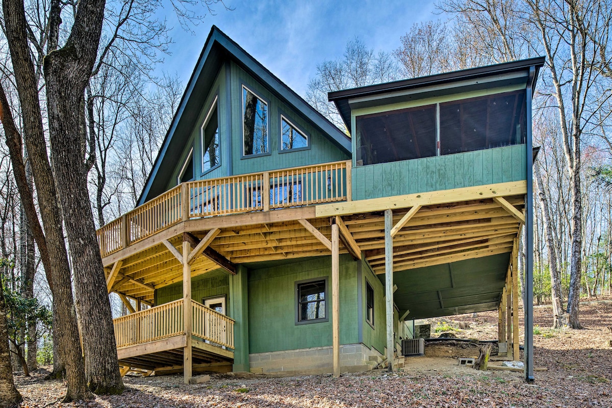 The multi-level cabin displays a charming green exterior surrounded by trees. A spacious deck wraps around the structure, featuring wooden railings. Large windows provide natural light to the interior. A screened porch is visible on the upper level, offering outdoor relaxation options.