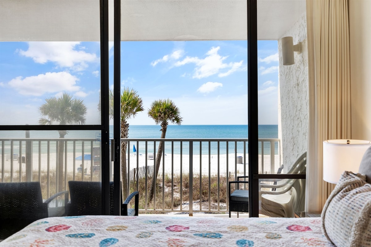 A beachfront view is presented from the sliding glass doors, showcasing the sandy shore and vibrant turquoise waters of the Gulf. Palm trees line the coastline, and a comfortable chair is positioned on the balcony, inviting relaxation in a tranquil setting.