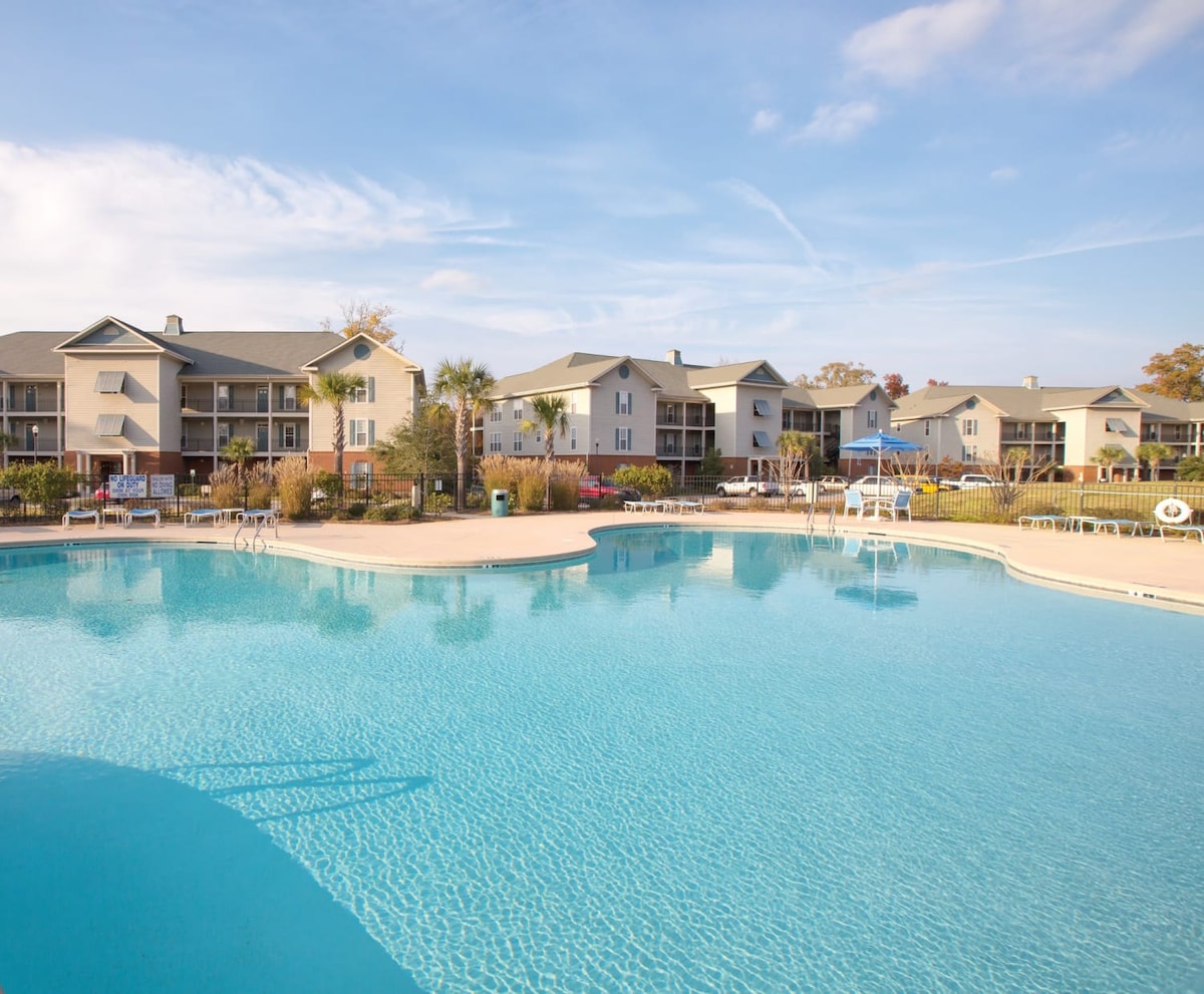 An outdoor swimming pool is depicted with clear blue water, surrounded by lounge chairs and landscaped gardens. The resort buildings are visible in the background, featuring balconies and a variety of exterior colors under a bright sky.