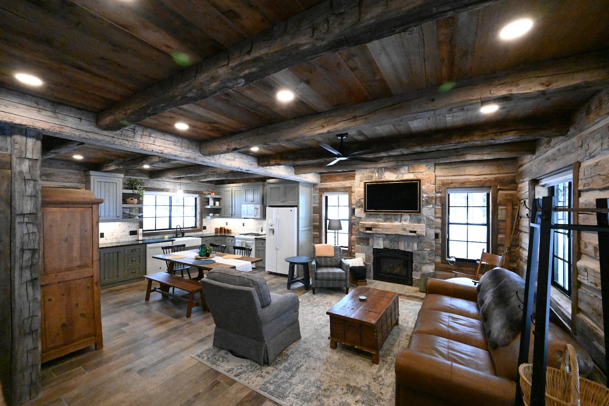 The living area features a blend of rustic and modern elements, with exposed wooden beams and stone accents. A comfortable seating arrangement includes a gray armchair and a brown leather sofa, facing a flat-screen TV and a cozy fireplace, all illuminated by warm overhead lighting.