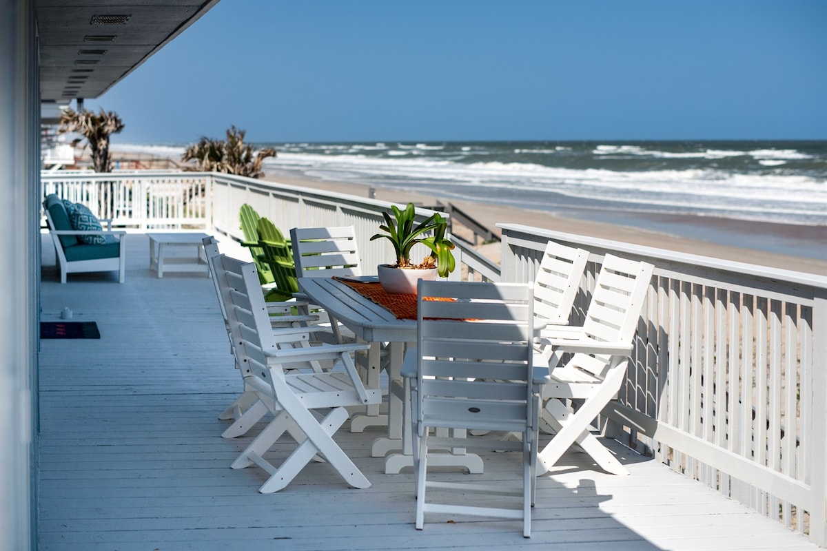 An expansive oceanfront deck features a large dining table surrounded by white chairs, inviting al fresco meals. Coastal views stretch across the sandy beach and waves, while accent chairs in vibrant colors add to the outdoor space's charm.