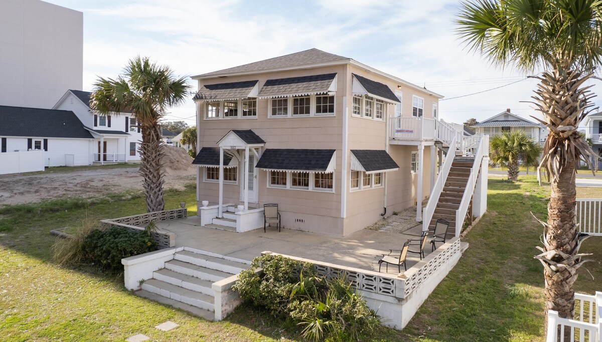 The exterior of a two-story house is visible, showcasing a pale beige façade with large windows. A welcoming patio area features several chairs arranged neatly, surrounded by palm trees and greenery. A staircase leads to the upper level, enhancing access and inviting relaxation.
