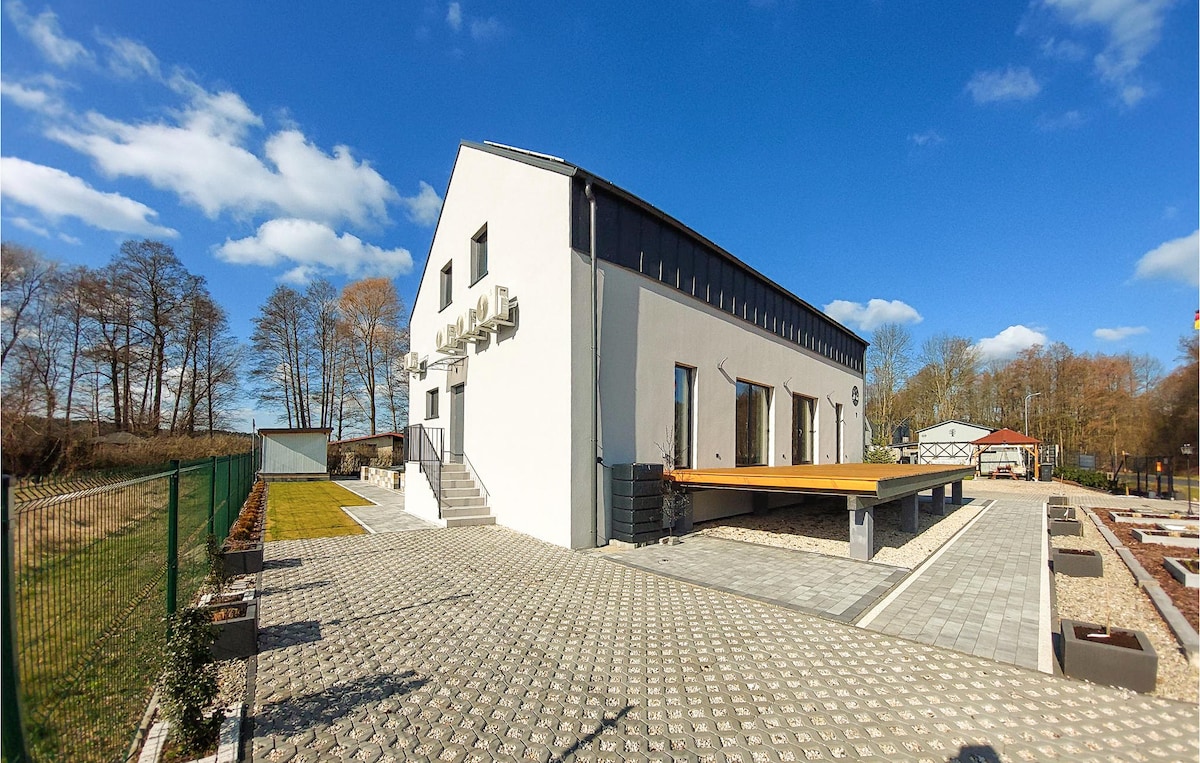 The exterior of the vacation home is presented, featuring a modern design with a white facade and large windows. A spacious wooden deck extends from the building, surrounded by a landscaped area with gravel paths. A grassy area is visible in the background with trees and open space.