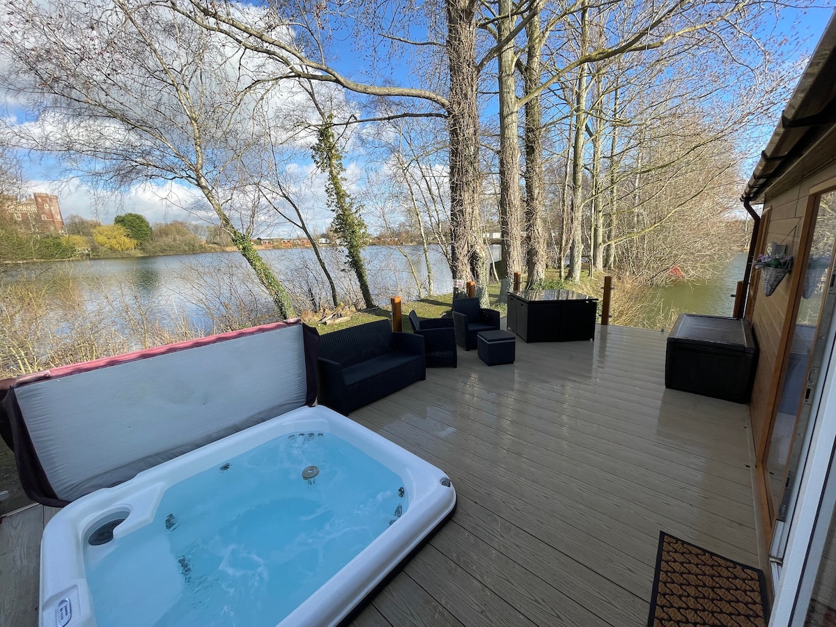 A spacious veranda overlooks Rudd Lake, featuring a sunken hot tub alongside comfortable seating. Trees provide a natural backdrop, enhancing the serene atmosphere by the water. The expansive decking invites relaxation and offers a view of the tranquil surroundings.