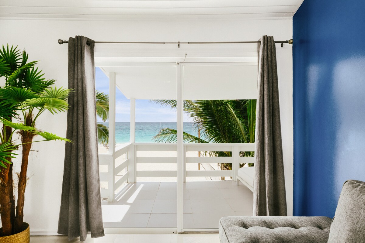 Floor-to-ceiling sliding glass doors open to a private balcony with ocean views, framed by lush palm trees. A cozy seating area, designed for relaxation, is complemented by a gray sofa and stylish drapes that softly filter the natural light.
