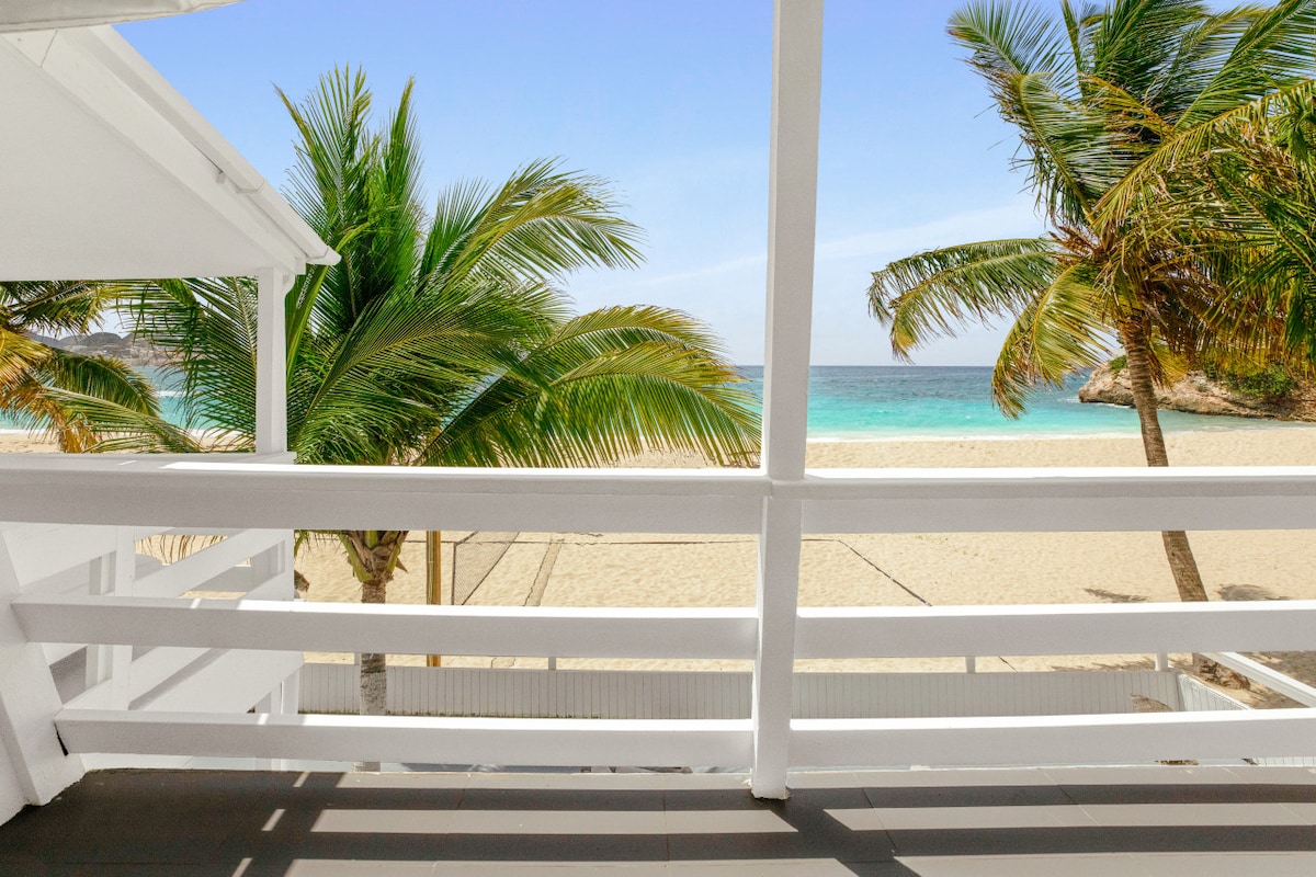 A view from a balcony showcases swaying palm trees with a backdrop of soft sandy beach and the turquoise sea. The horizon meets a clear sky, enhancing the serene coastal setting.