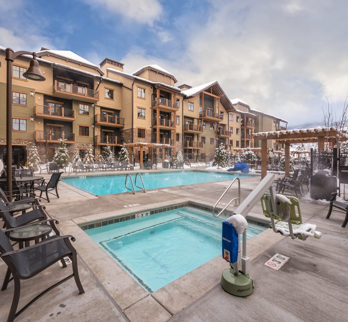 An outdoor swimming pool area is surrounded by snow-capped buildings, featuring seating options and a hot tub. The paved deck offers easy access to the pool, while snowy trees add a serene touch to the landscape.
