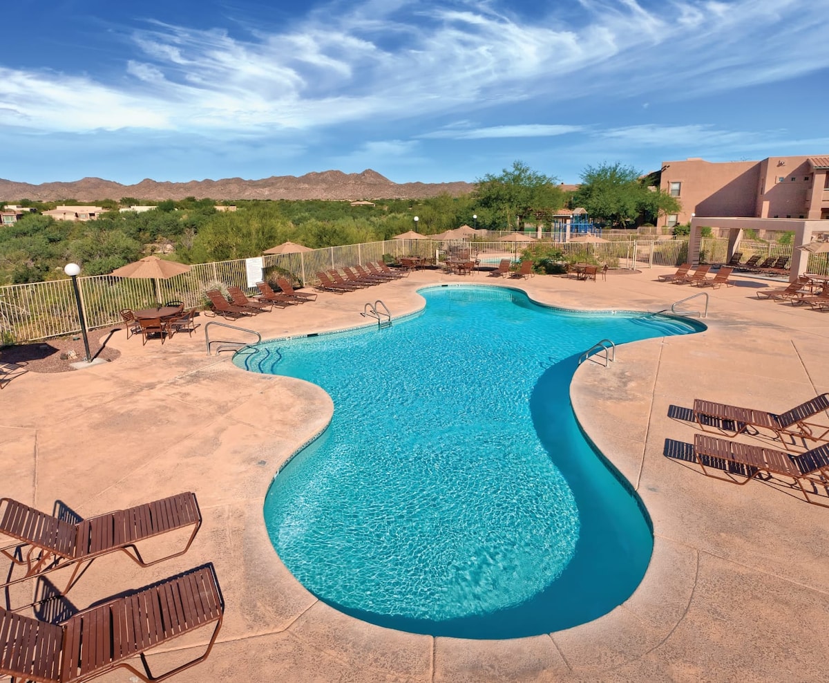 An outdoor swimming pool features a unique shape with a stunning turquoise hue. Surrounding the pool are numerous lounge chairs arranged neatly on a sun-drenched patio. Lush desert landscape and distant mountains provide a scenic backdrop under a clear blue sky.