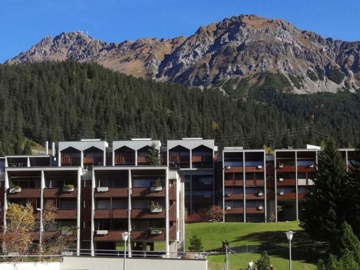 The modern apartment building is nestled against a backdrop of majestic mountains. The structure features multiple levels with balconies, while a lush green area is visible in the foreground, enhancing the natural setting. Clear blue skies contribute to the serene atmosphere.