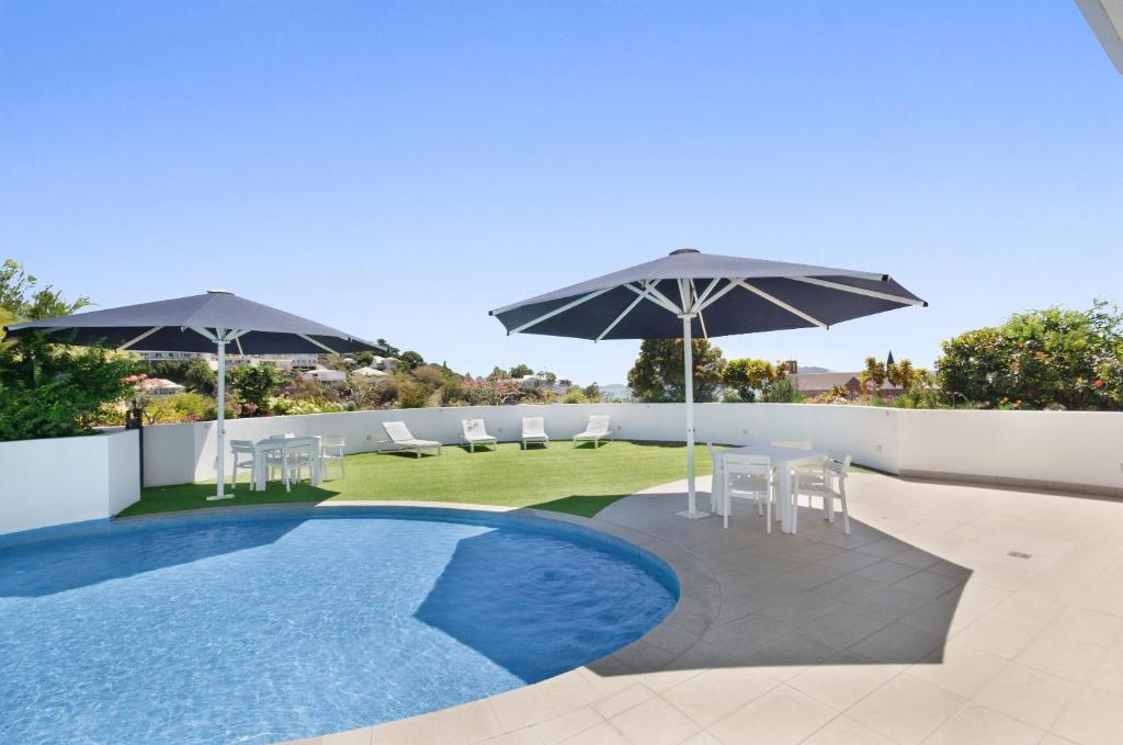 A rooftop area features a refreshing pool surrounded by green grass. Two large umbrellas provide shade over a selection of white lounge chairs and a dining table with matching chairs, inviting relaxation and outdoor enjoyment.