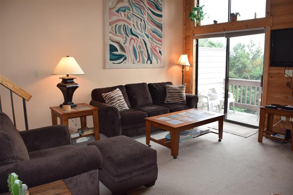 A comfortable living room features a dark sectional sofa and two wooden side tables. A coffee table sits in the center, and a flat-screen TV is mounted on the wall. Large glass doors provide access to a deck area and allow natural light to fill the space.
