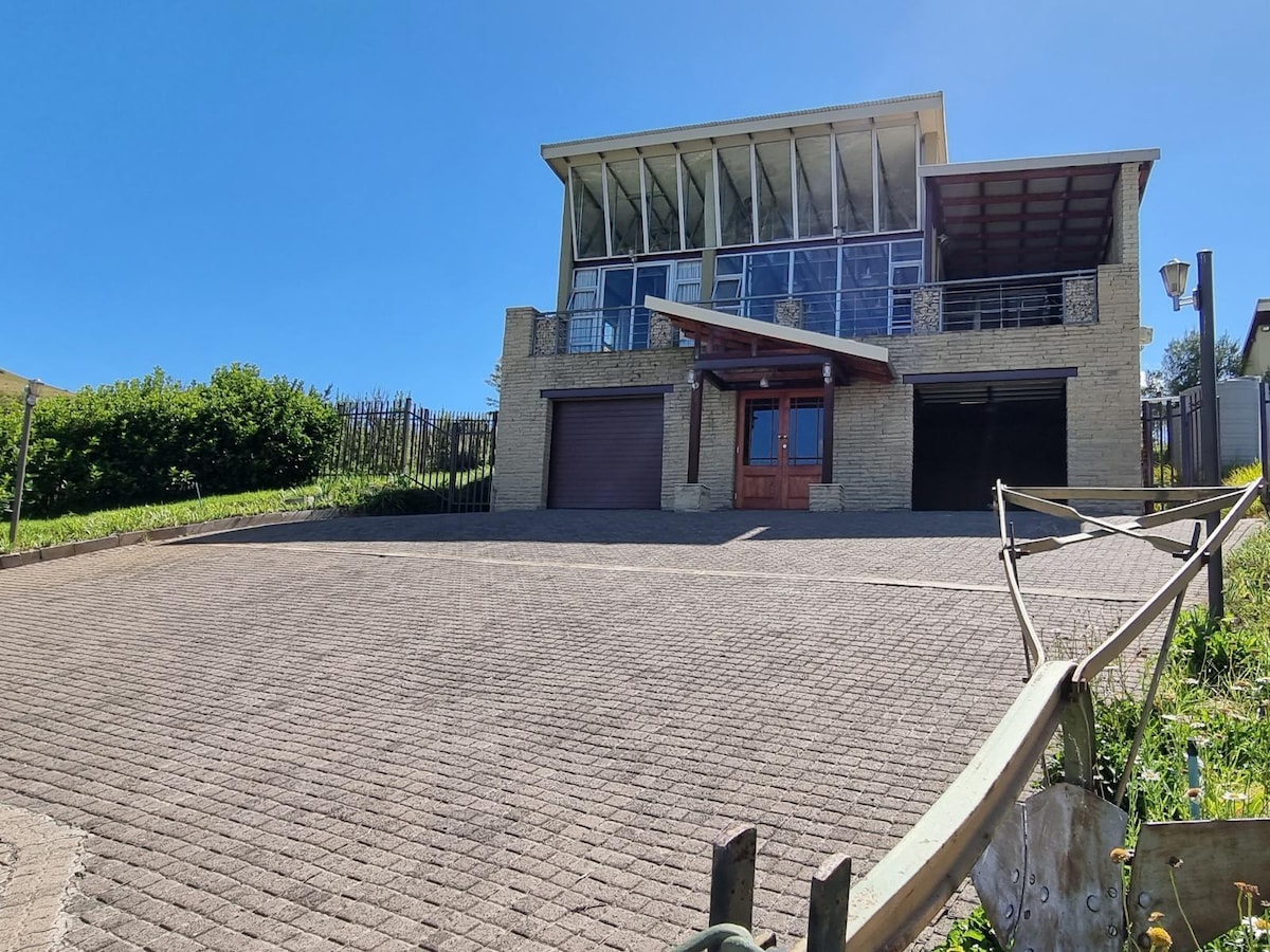 The exterior of a modern two-story house is shown, featuring large windows that provide ample natural light. The garage door is visible, alongside a covered entryway. The driveway is paved, and green vegetation surrounds the property, contributing to a peaceful atmosphere.