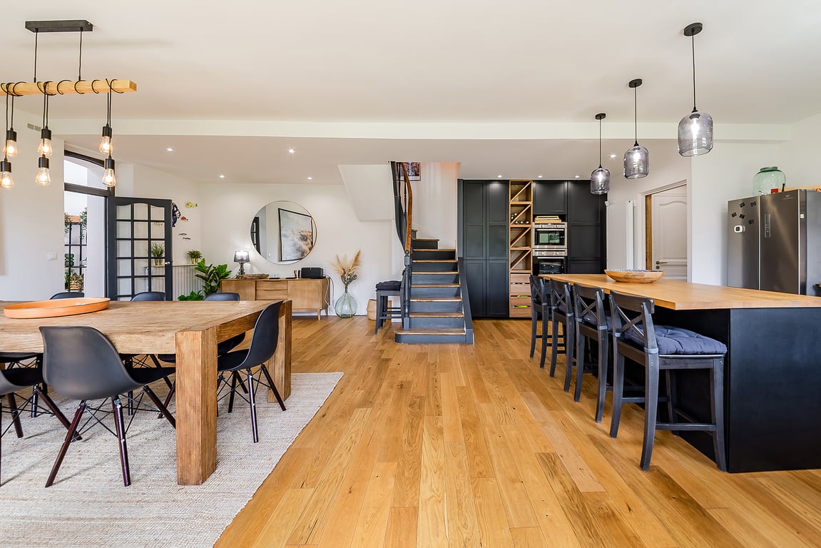 An open-concept dining and kitchen area features a large wooden dining table surrounded by black chairs. A central kitchen island with bar seating complements modern appliances and cabinetry. A staircase leads to the upper level, with natural light illuminating the space through large windows.
