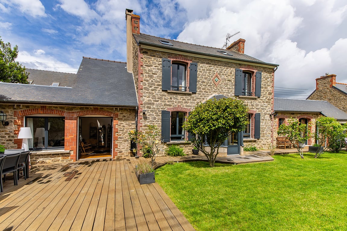 The exterior of the renovated house features a stone facade with blue shutters. A spacious wooden deck provides outdoor seating, surrounded by well-maintained grass and greenery. Large windows allow natural light to illuminate the interior spaces beyond.