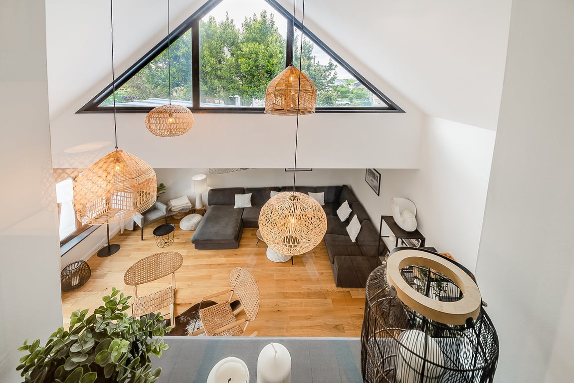 An airy living room features a mix of natural textures and modern furnishings. Large triangular windows invite light, illuminating a spacious seating area with a dark sofa. Hanging light fixtures made of woven materials add warmth, while an arrangement of tables and chairs offers options for relaxation.