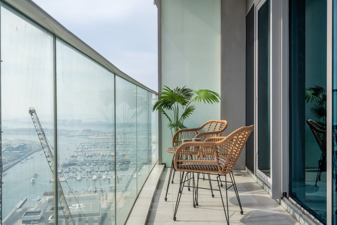 A modern balcony offers a view of the marina, featuring two stylish rattan chairs positioned beside a small potted palm. The space is enclosed by floor-to-ceiling glass panels that provide unobstructed views of the surroundings.