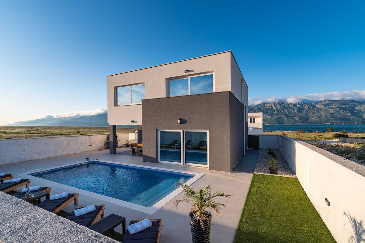 A modern villa with a two-story design features large windows and a private swimming pool in the foreground. A fenced courtyard surrounds the pool, with sun loungers arranged neatly. The backdrop showcases the stunning Velebit mountain range and a clear blue sky.