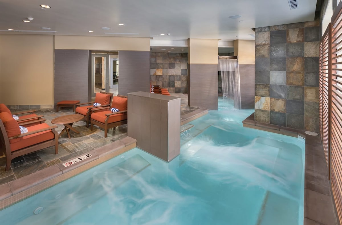 A relaxing spa area is presented with soft blue water flowing through hot tubs and jets. Comfortable orange lounge chairs are positioned around the space, alongside natural stone and tiled surfaces, creating a tranquil environment for guests.