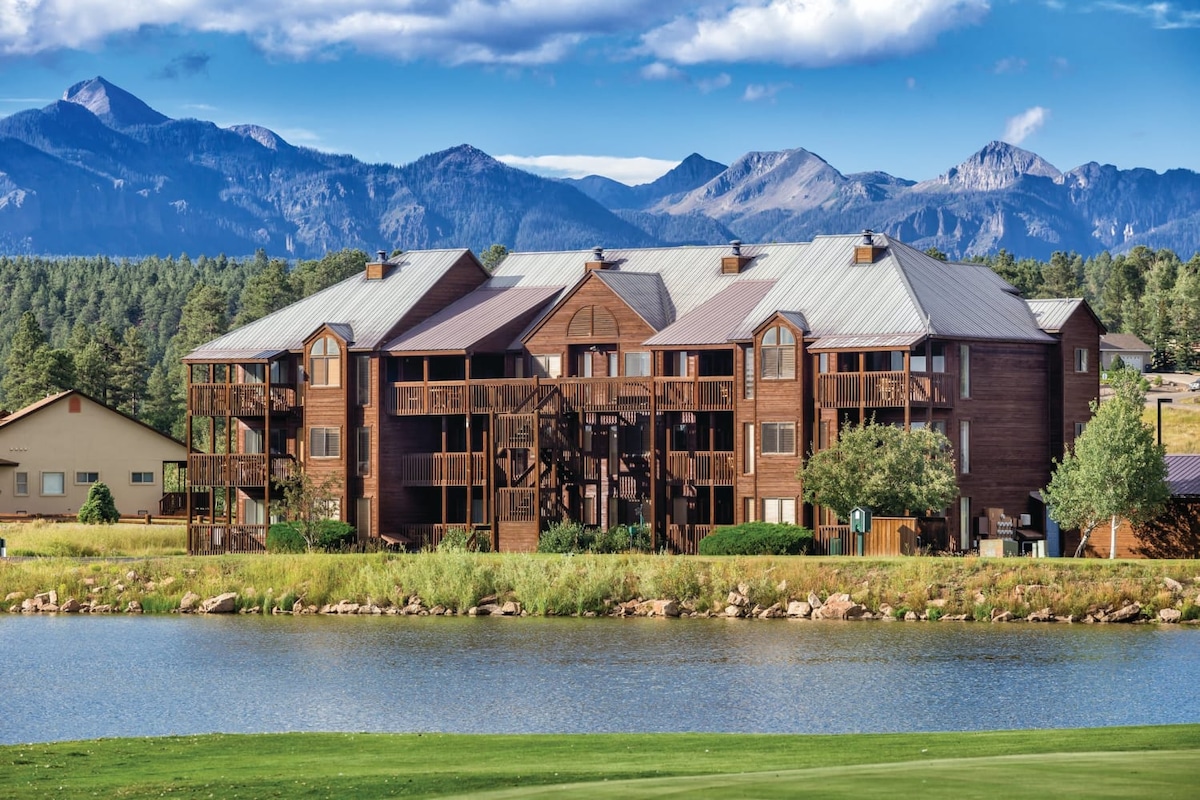 A multi-story building constructed with natural wood accents is situated beside a calm lake. Towering mountains form a picturesque backdrop, and lush greenery surrounds the property. Balconies enhance the outdoor appeal, providing potential views of the landscape.