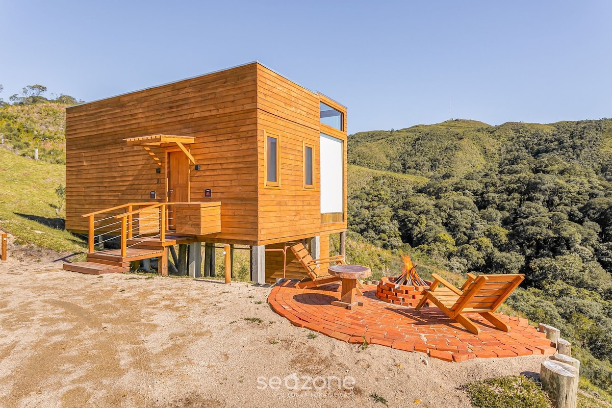A modern wooden cabin is set on a hillside, showcasing large windows that capture natural light. An outdoor seating area is arranged with lounge chairs and a fire pit, surrounded by lush green hills in the background. A clear blue sky enhances the serene atmosphere.
