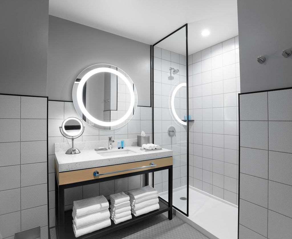 A modern bathroom features a clean design with white tiled walls and a walk-in shower. A round mirror with built-in lighting is mounted above a sleek vanity with a stone countertop. Fresh towels are neatly stacked on a shelf below the sink.