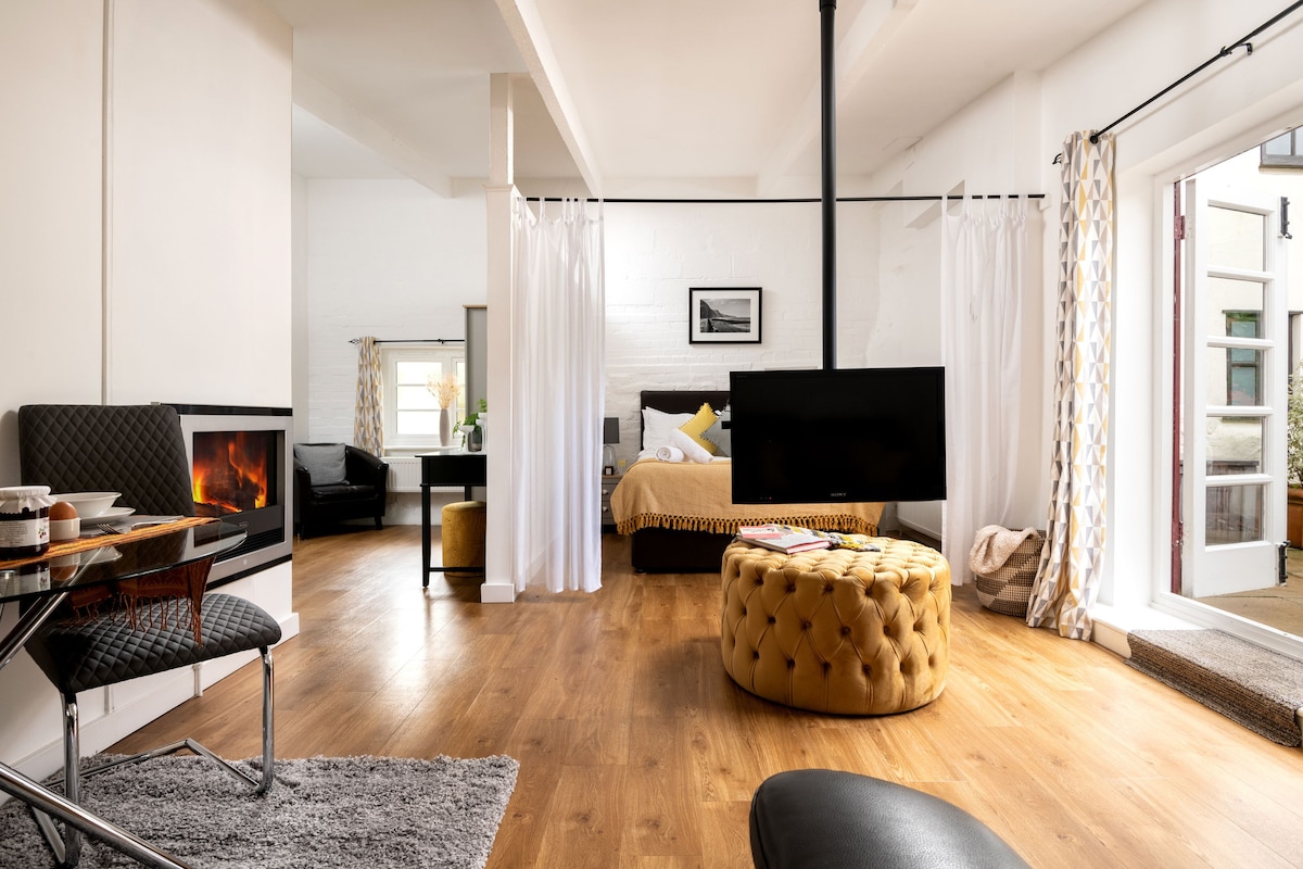 An open-plan living area is highlighted by a cozy electric wall fire and a stylish mounted TV. The space features wooden flooring, a comfortable bed visible in the background, and light flowing through large windows draped with sheer curtains. A circular ottoman adds a touch of elegance.