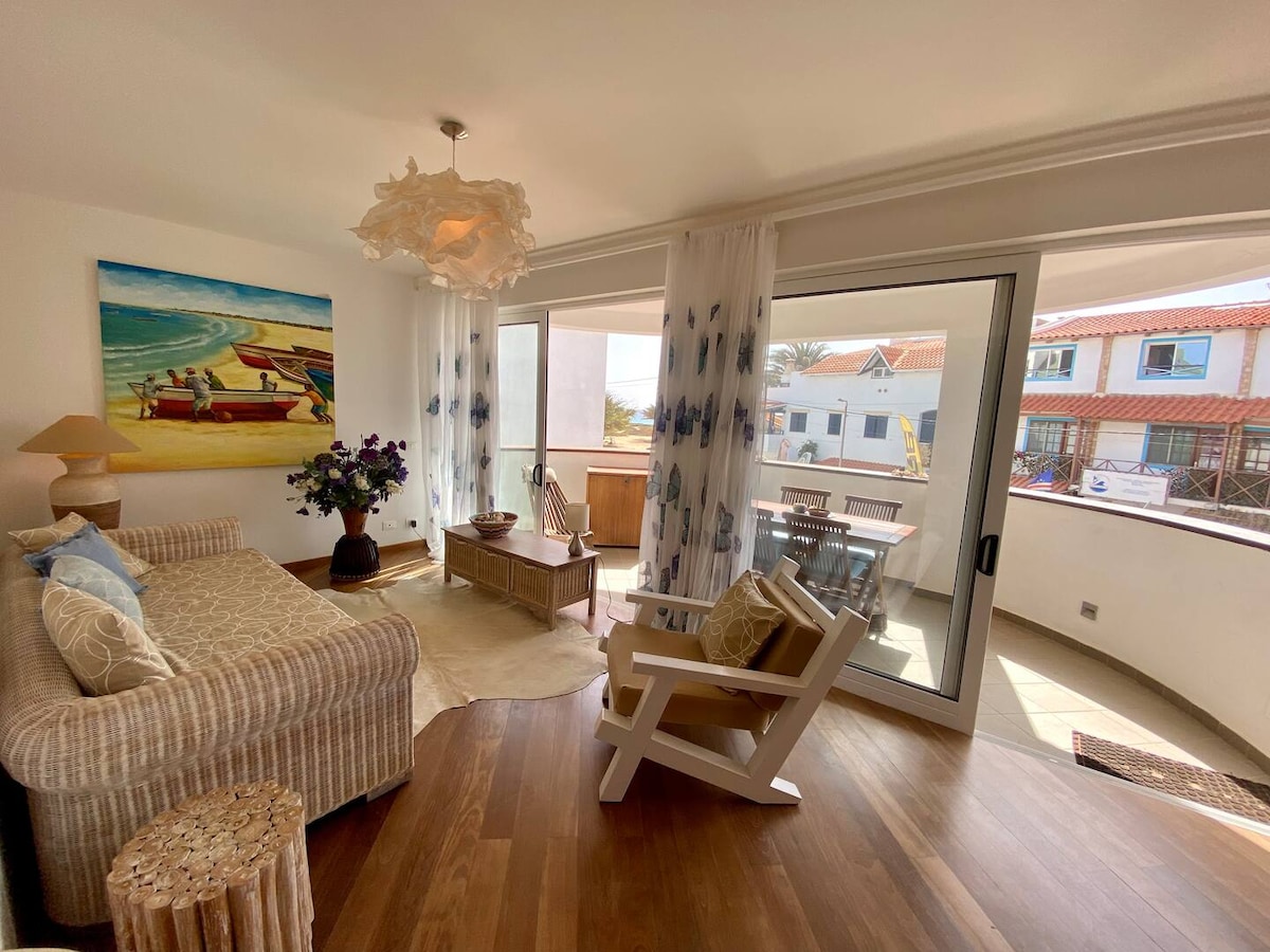 A sunny living area features a comfortable sofa paired with an armchair, positioned near large glass doors that open to a private terrace. Light flowing through sheer curtains creates an airy feel, and decorative elements include a light fixture and a small side table.