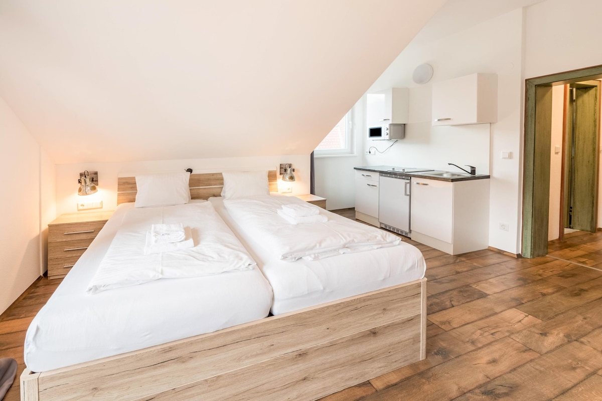 The bedroom features a spacious arrangement of two beds with crisp white linens. Soft bedside lighting provides a gentle glow. An adjoining kitchen area is visible, equipped with modern amenities and cabinetry, along with a separate entryway leading to other parts of the apartment.