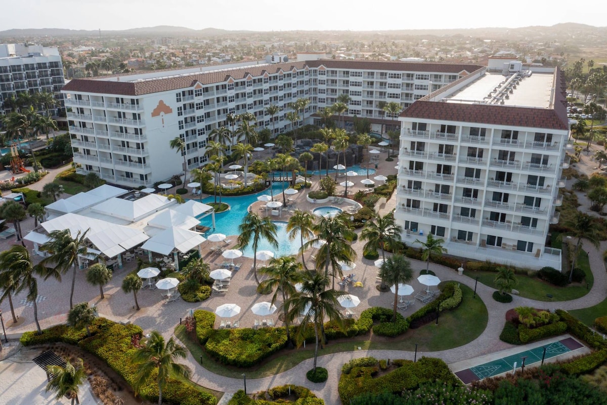 An aerial view captures the resort's layout, showcasing multiple buildings surrounded by landscaped gardens. The large outdoor pool is visible, complemented by seating and umbrellas. Palm trees add to the tropical ambiance, and paths meander through the grounds, providing access to various areas.
