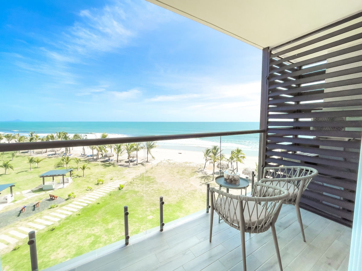 A private balcony offers a view of the beach and ocean beyond. Two stylish chairs are positioned around a small table, creating a cozy seating area. Lush green grass and palm trees frame the beach, providing a serene outdoor atmosphere.