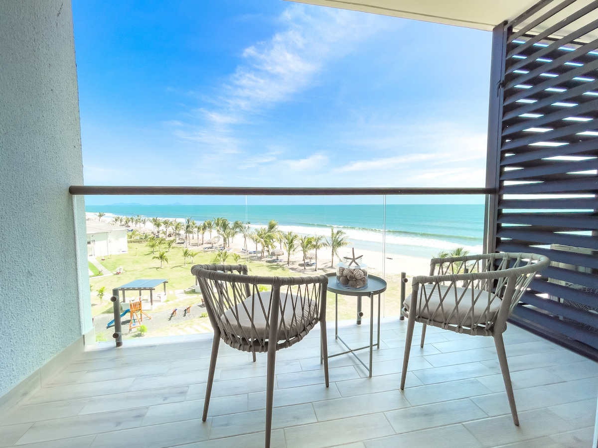 A private balcony features two lightweight chairs and a small table. The view overlooks the beach and ocean, with palm trees lining the shore. Natural light enhances the open space, creating a relaxed atmosphere for enjoying the coastal scenery.
