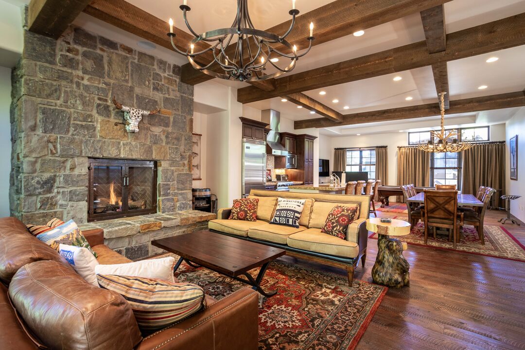 A spacious living area features a stone fireplace, wooden beams overhead, and comfortable seating. A large coffee table sits on a textured rug. In the background, a dining table accommodates multiple guests, with warm lighting creating a welcoming space.