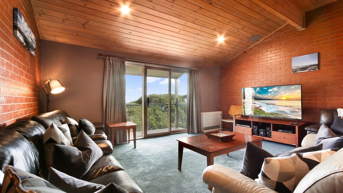 A living area features a cozy arrangement of dark leather sofas and a wooden coffee table. Large glass doors provide access to a scenic view outside. A flat-screen TV is positioned on a media unit against a brick wall, while soft lighting enhances the warm ambiance.