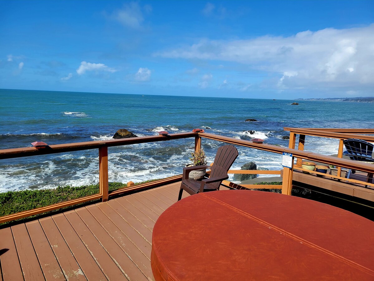 A spacious deck presents stunning views of the Pacific Ocean, with gentle waves lapping against rocky outcrops. A full-size hot tub is positioned beside an Adirondack chair, inviting relaxation in front of the expansive water vista under a bright blue sky.