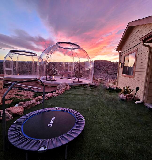 Two clear glamping domes are positioned on the property, surrounded by rocky terrain and a vibrant sunset sky. A trampoline is set on the grassy area in the foreground, enhancing the outdoor recreational space near the home.